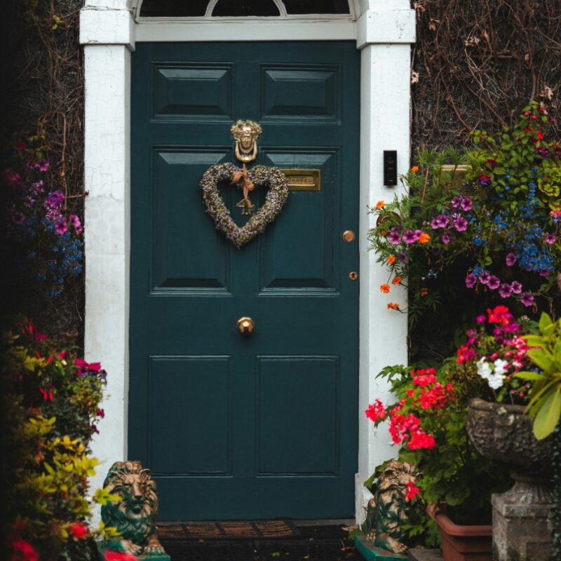 A dark teal front door with white trim and an arched fanlight window, decorated with a heart-shaped floral wreath and brass hardware. Colorful flowering plants, vines, and two small lion statues flank the entrance.