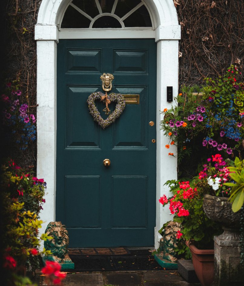 A dark teal front door with white trim and an arched fanlight window, decorated with a heart-shaped floral wreath and brass hardware. Colorful flowering plants, vines, and two small lion statues flank the entrance.