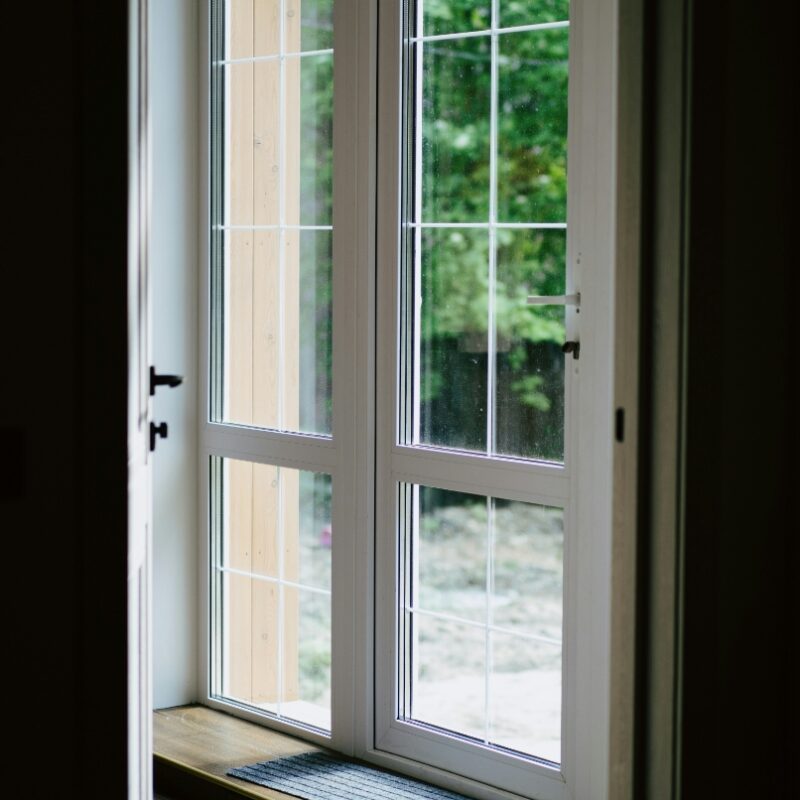 A white-framed glass patio door with multiple window panes, viewed from inside and opening onto a green outdoor area. A dark doormat rests on the wooden threshold.