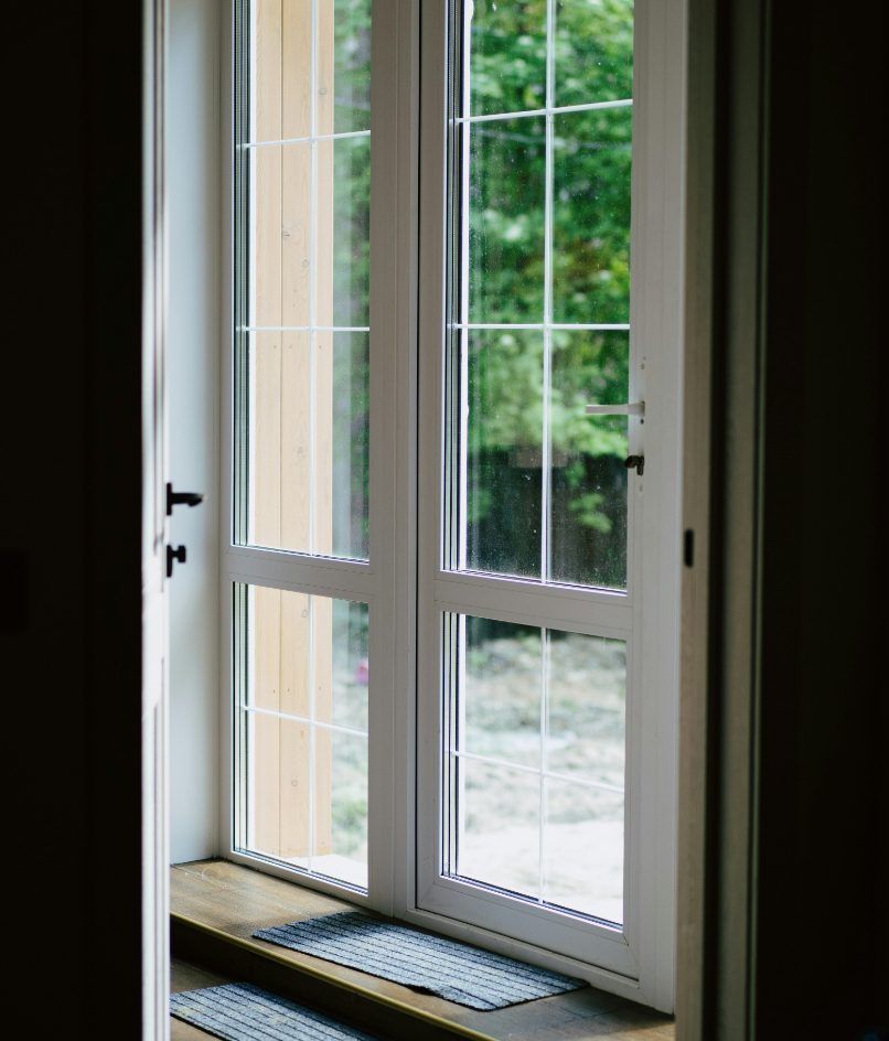 A white-framed glass patio door with multiple window panes, viewed from inside and opening onto a green outdoor area. A dark doormat rests on the wooden threshold.